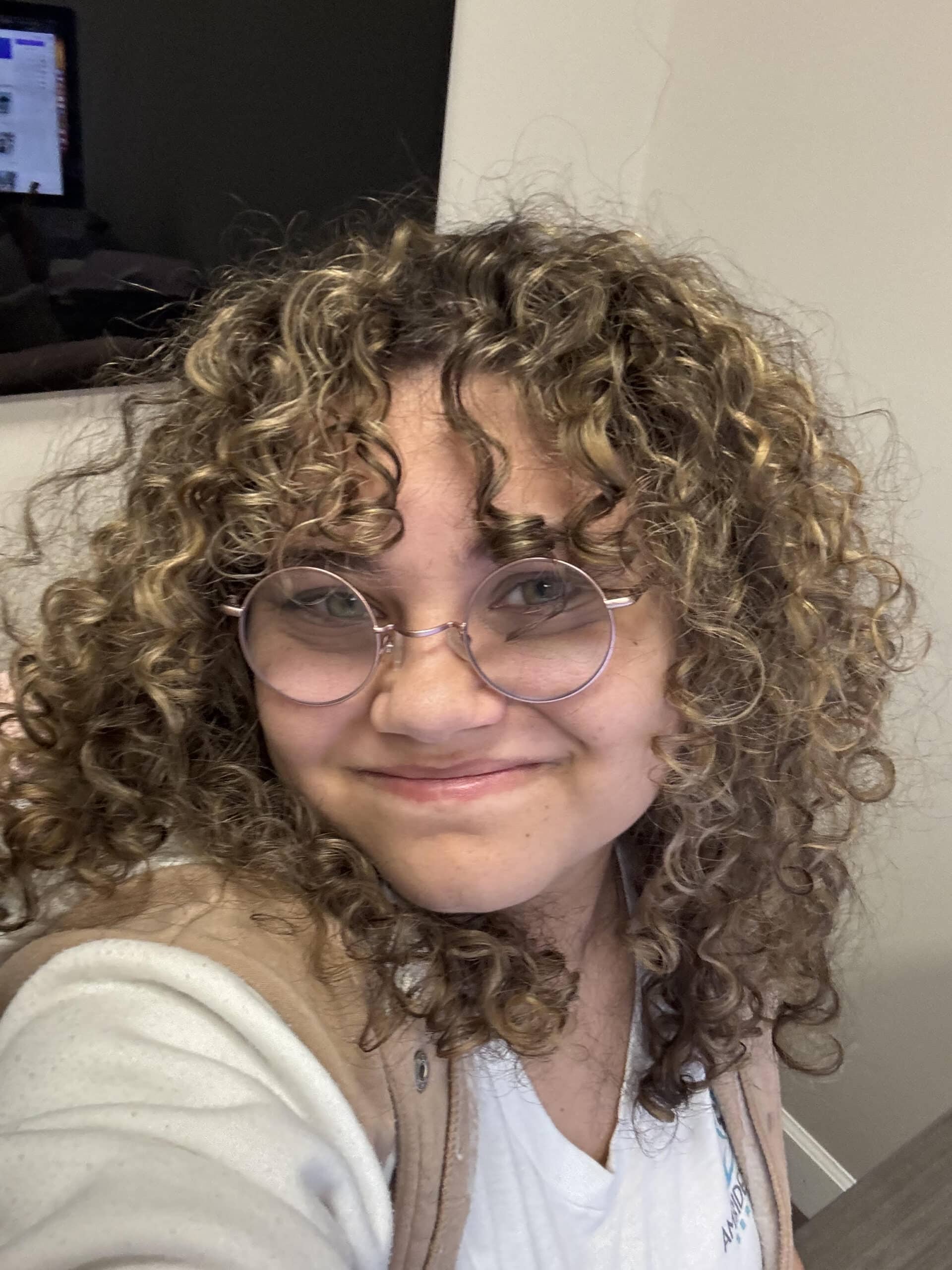 Amidst the glow of ambition's fiery rise, a person with curly hair and round glasses smiles at the camera, embodying the rebirth of creativity through their ambidextrous skills. Clad in a light-colored top and vest, they emerge from behind the screen's digital ashes, where a computer monitor on a desk flickers with hints of their versatile services.